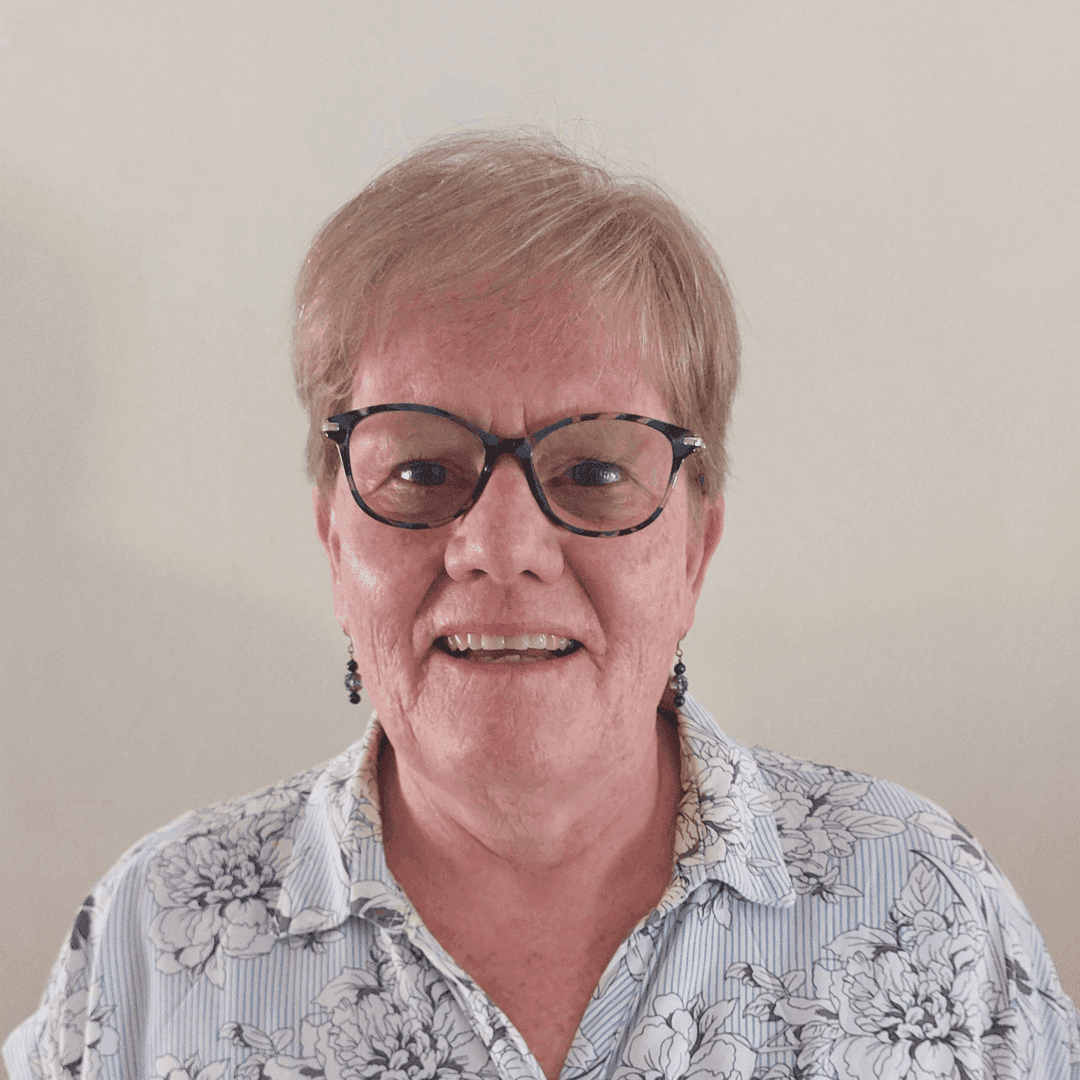 Smiling elderly person with glasses, short hair, and a patterned shirt against a plain background. - Home Instead
