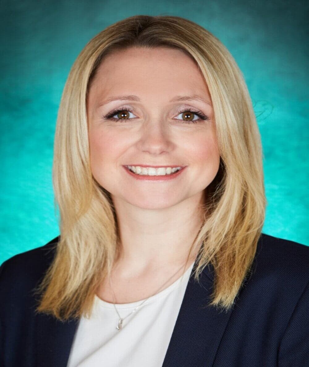 A woman with shoulder-length blonde hair smiles, wearing a dark blazer over a white top against a teal background. - Home Instead
