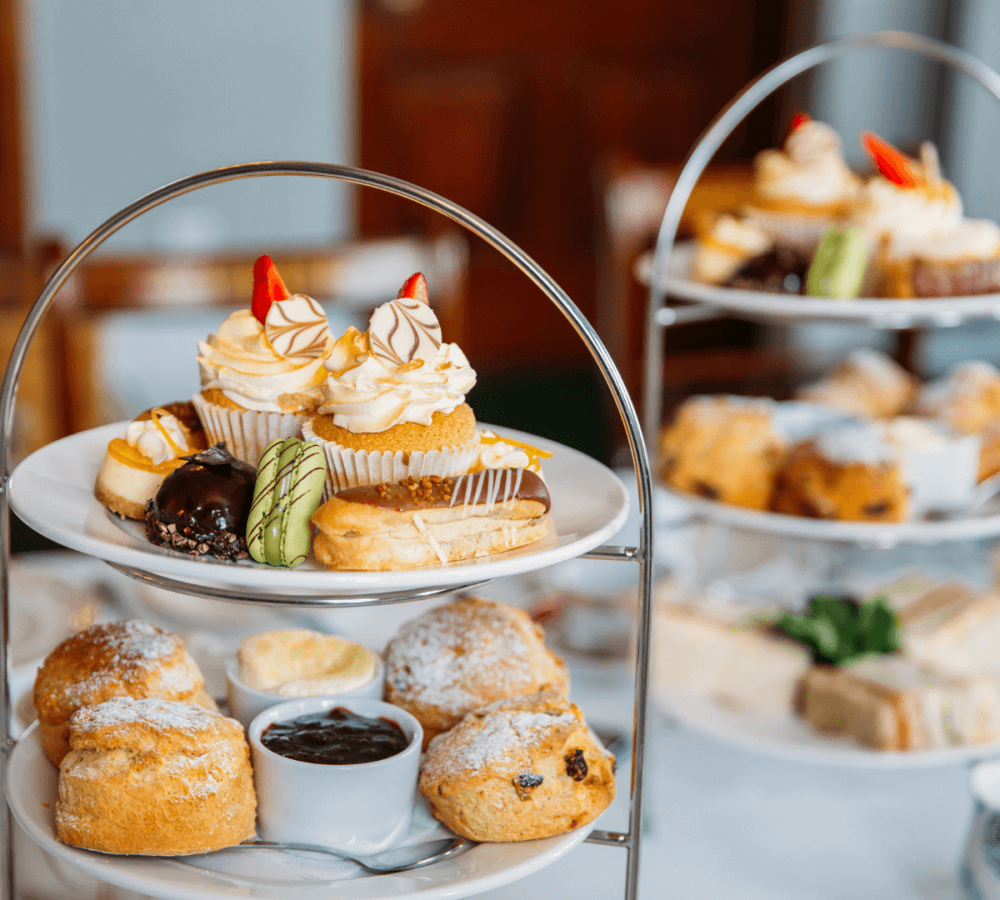 Afternoon tea setup with tiered trays featuring pastries, scones with clotted cream and jam, and various cakes. - Home Instead