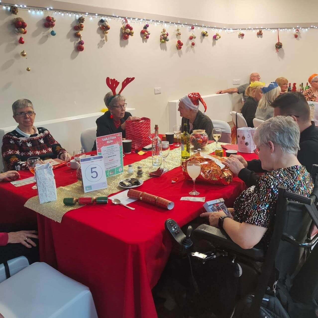 A group of people in festive attire sit around a decorated table at a holiday gathering, enjoying food and drinks. - Home Instead
