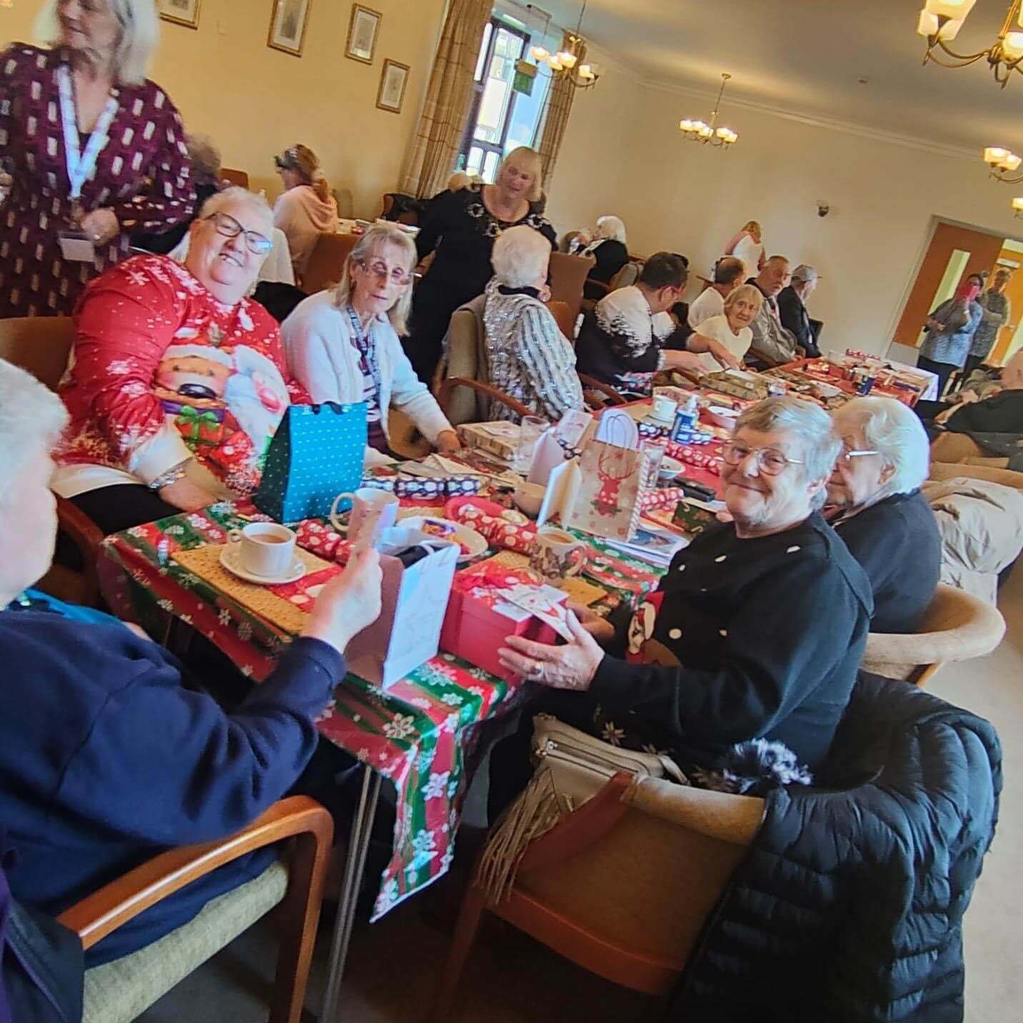 A group of people enjoying a festive holiday party at a communal table, surrounded by decorations and gifts. - Home Instead