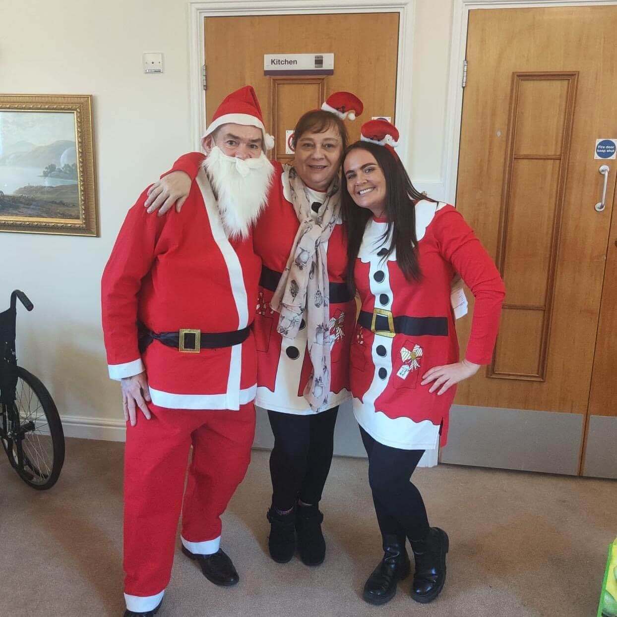 Two people dressed as Santa and a woman in a festive scarf stand together, smiling in a room with a sign labeled "Kitchen. - Home Instead