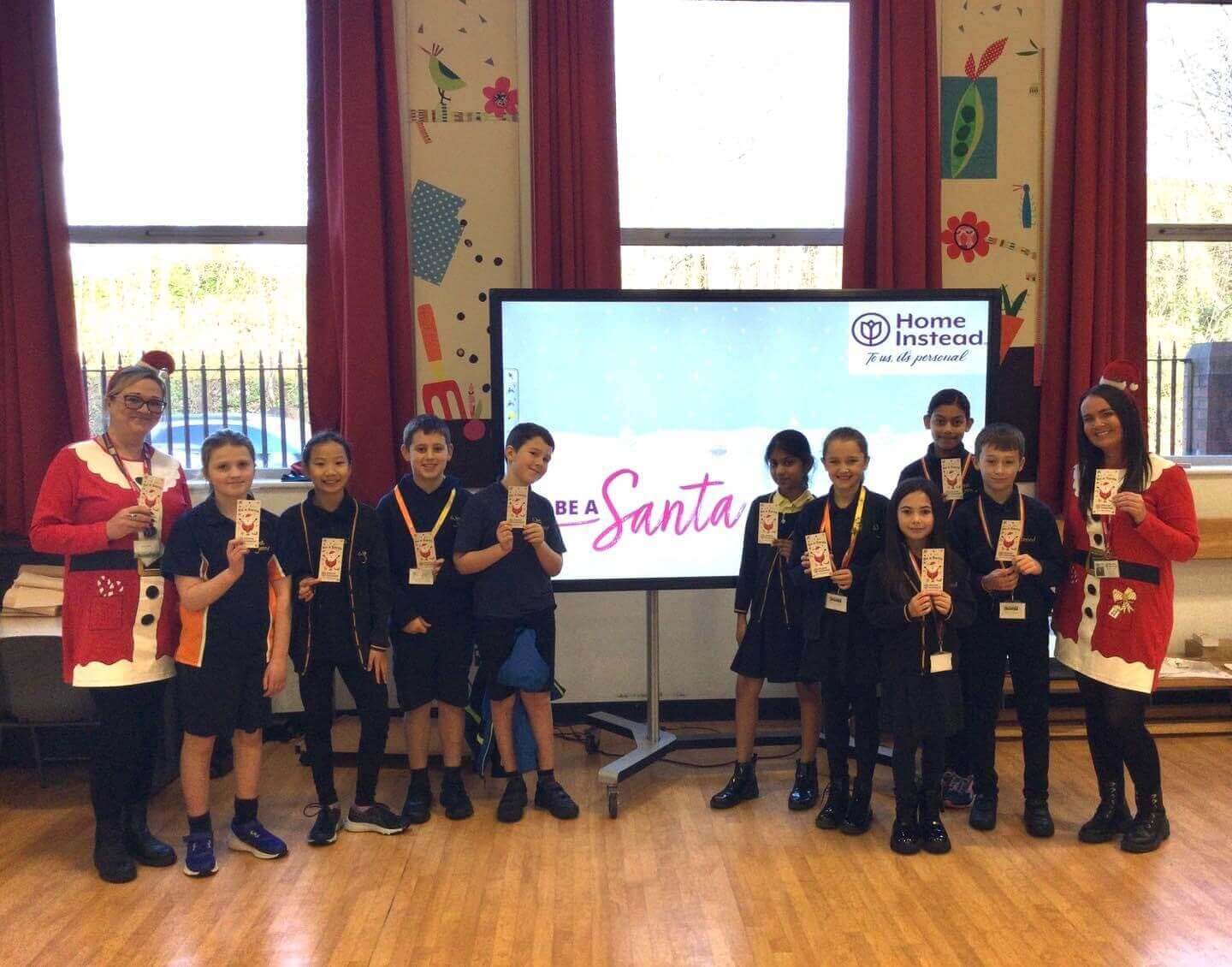 A group of children and two adults stand in front of a screen displaying "Be a Santa" in a decorated room. - Home Instead