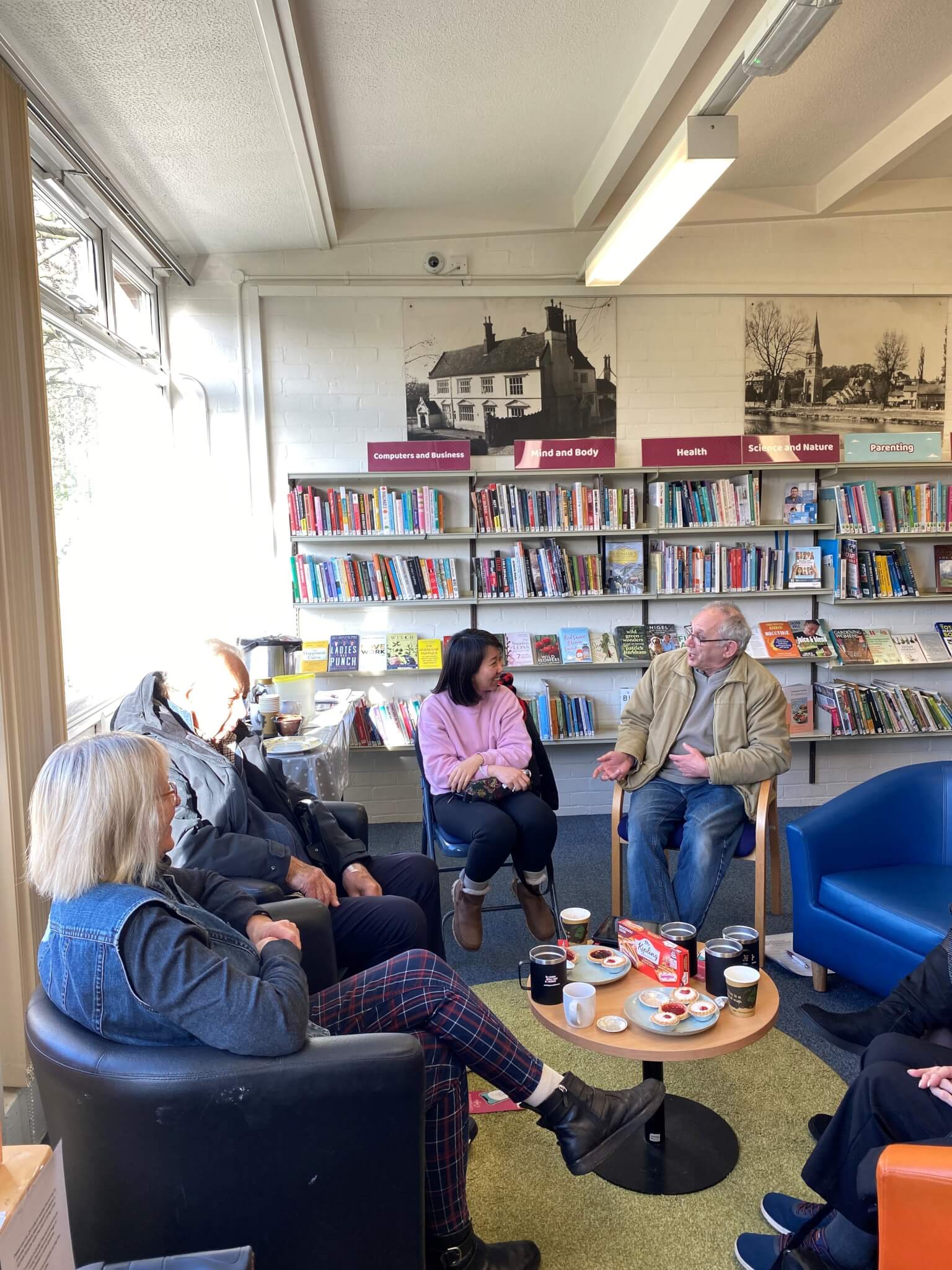 A small group of people sitting in a library, engaged in conversation, with coffee cups and snacks on a table. - Home Instead