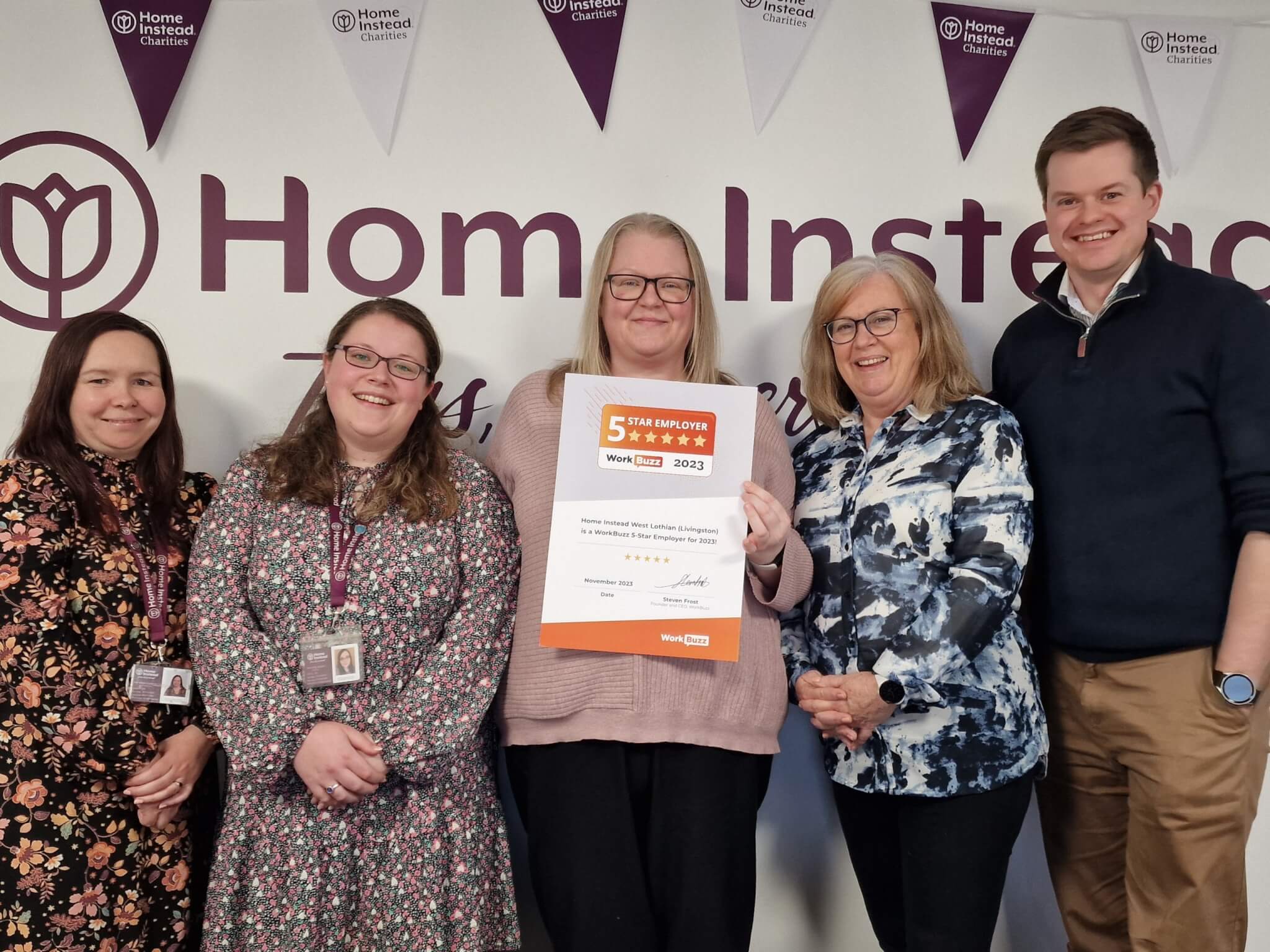 Five people smiling and holding a certificate in front of a "Home Instead" banner with celebratory flags. - Home Instead