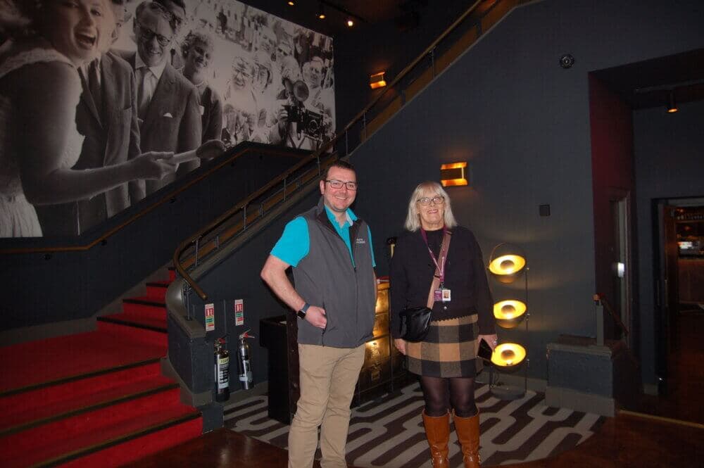 Two people standing inside a modern venue near a staircase. They are both smiling and posing for the camera. - Home Instead