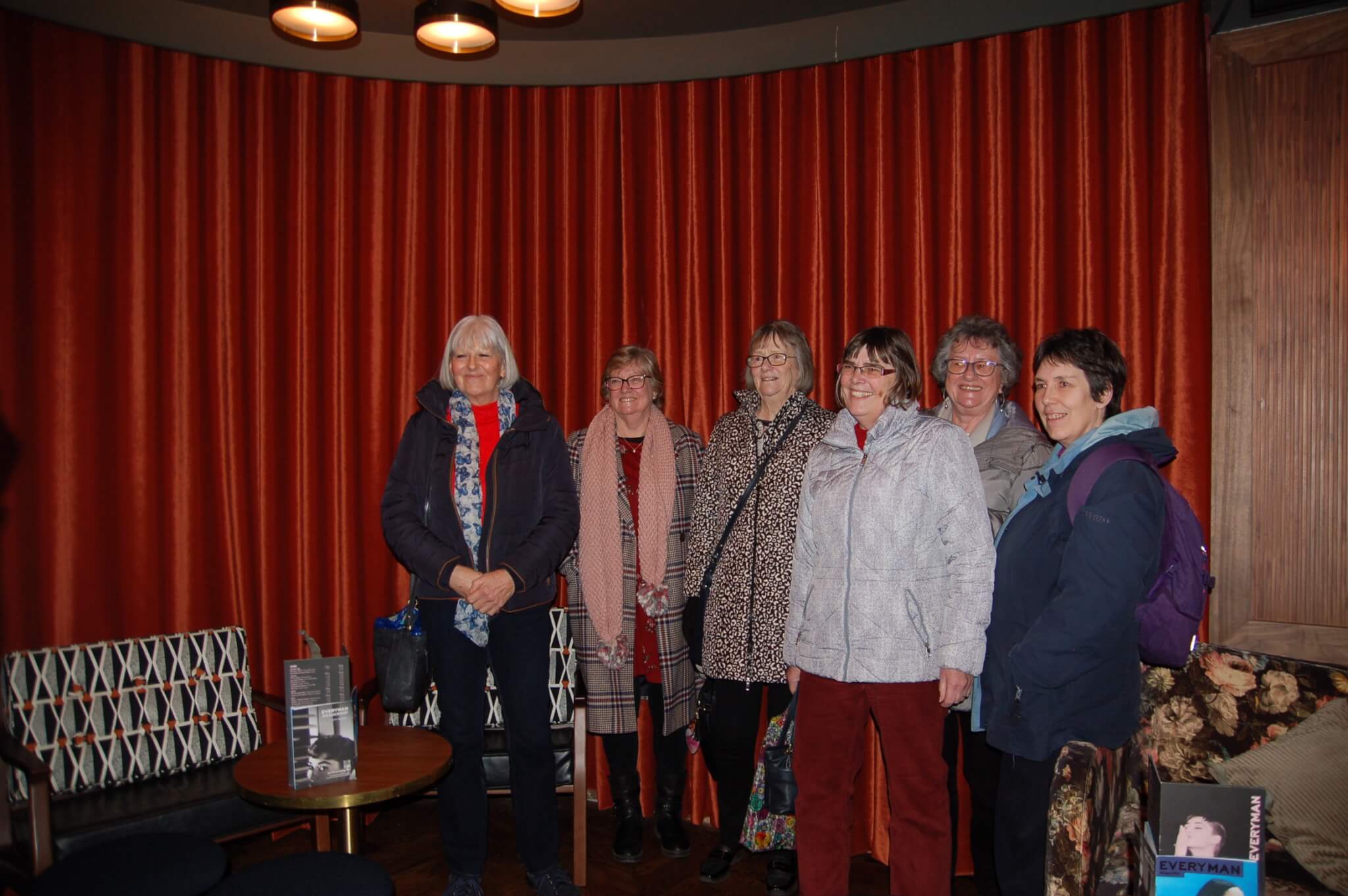 Six elderly women standing together in front of a red curtain, smiling at the camera. - Home Instead