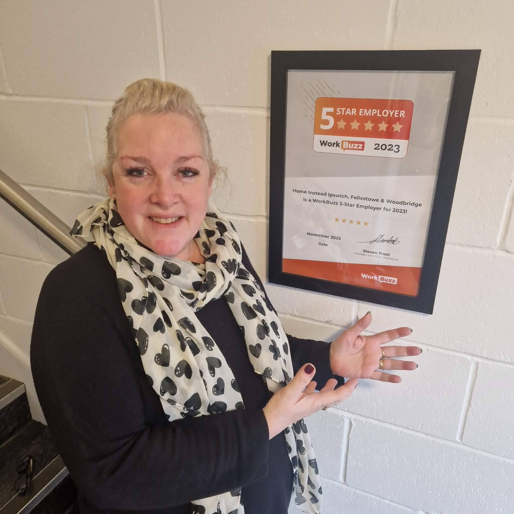 A smiling woman points to a framed “5 Star Employer” certificate from WorkBuzz, dated 2023, mounted on a wall. - Home Instead