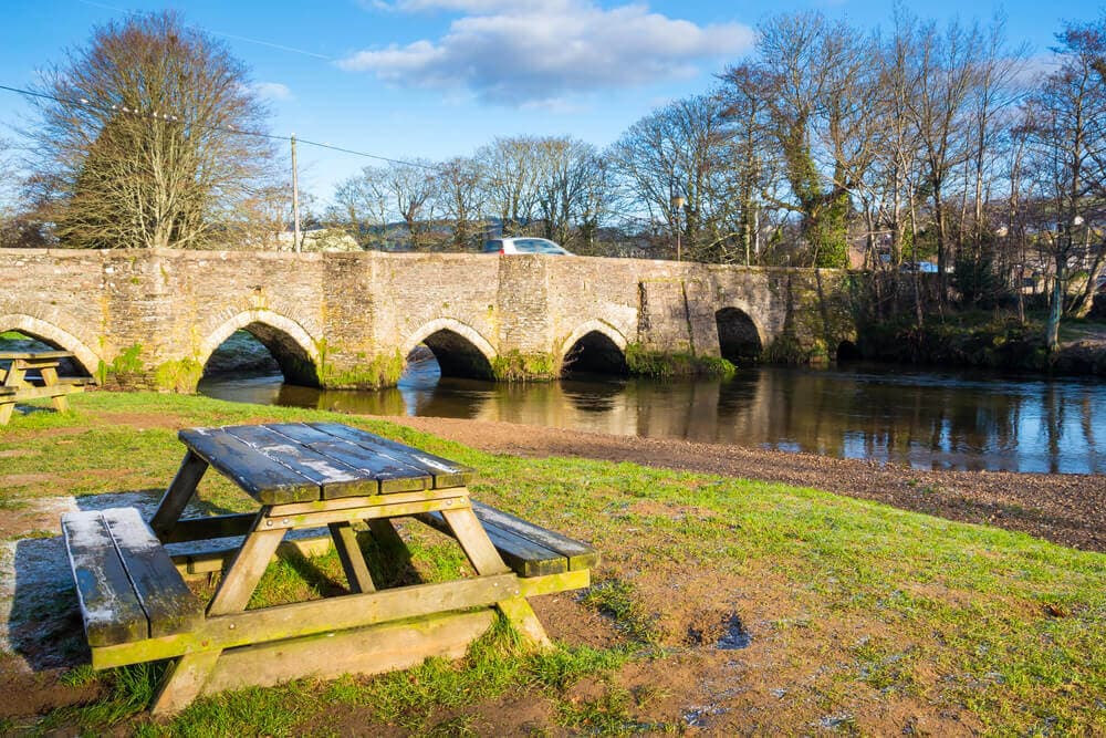 A wooden picnic table near a calm river with an old arched stone bridge in the background on a sunny day. - Home Instead