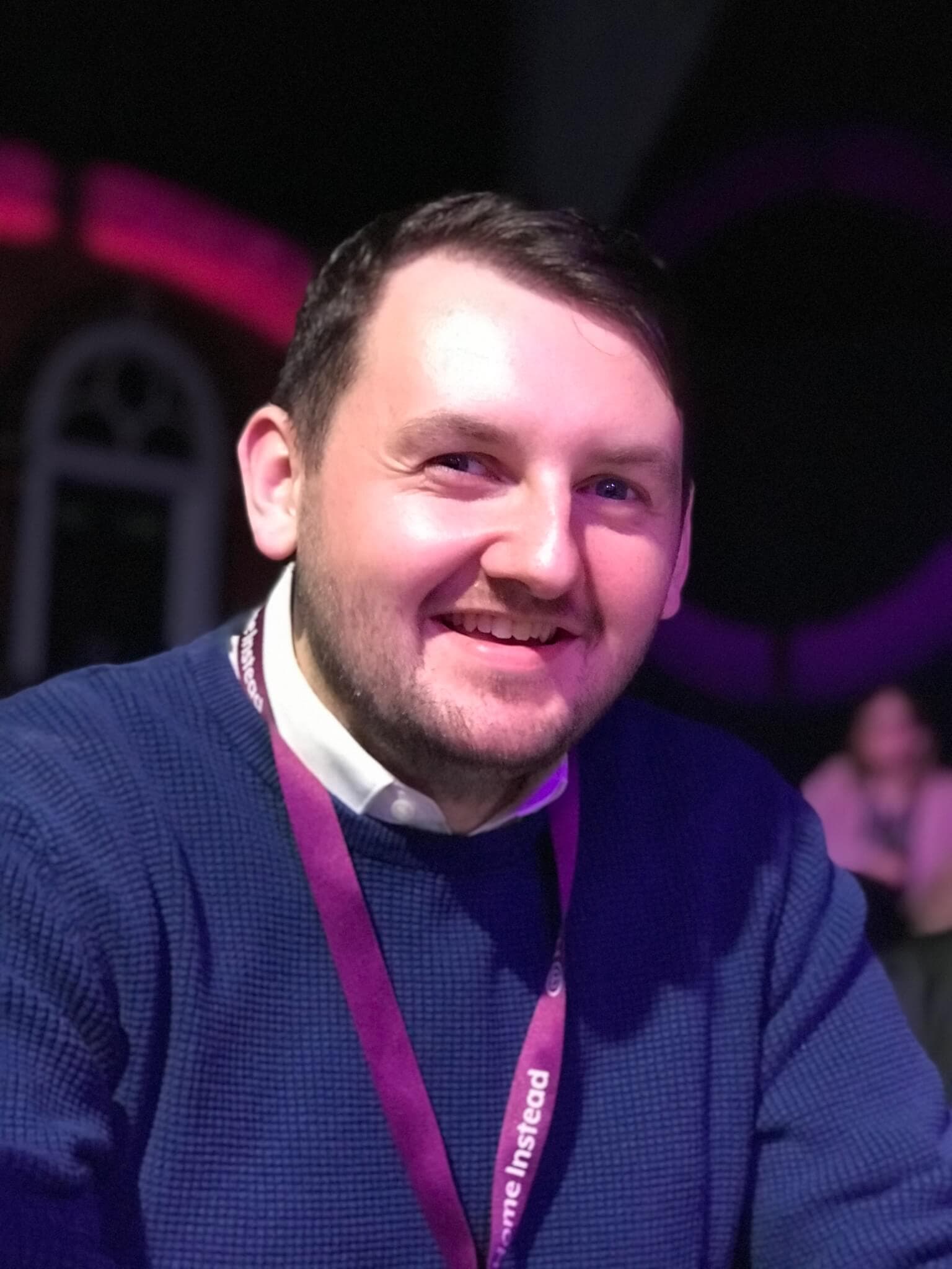 A man with short hair and a beard, wearing a blue sweater and lanyard, smiles while seated in a dimly lit area. - Home Instead