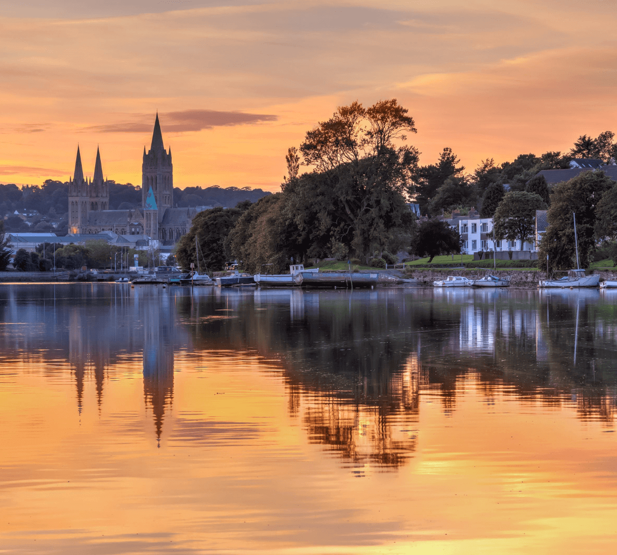 Sunset over a calm river with a cathedral and trees reflecting in the water, boats moored along the shore. - Home Instead
