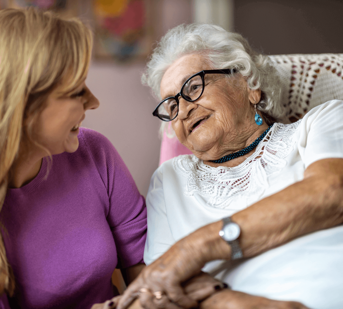 Young woman and elderly woman smiling warmly at each other while holding hands in a comforting gesture. - Home Instead