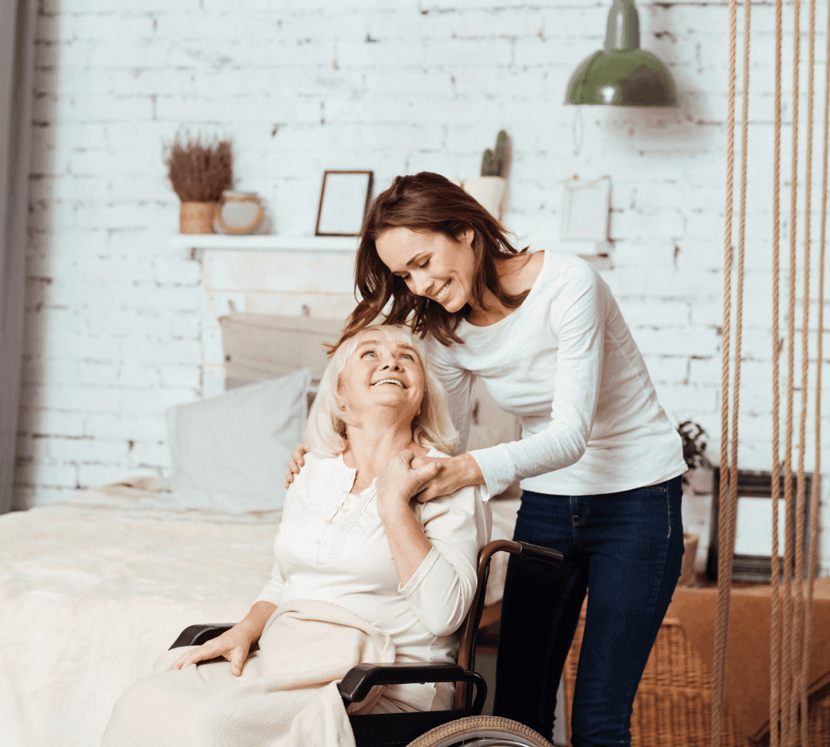 A smiling woman in a wheelchair is embraced by a standing woman in a bright, cozy room. - Home Instead