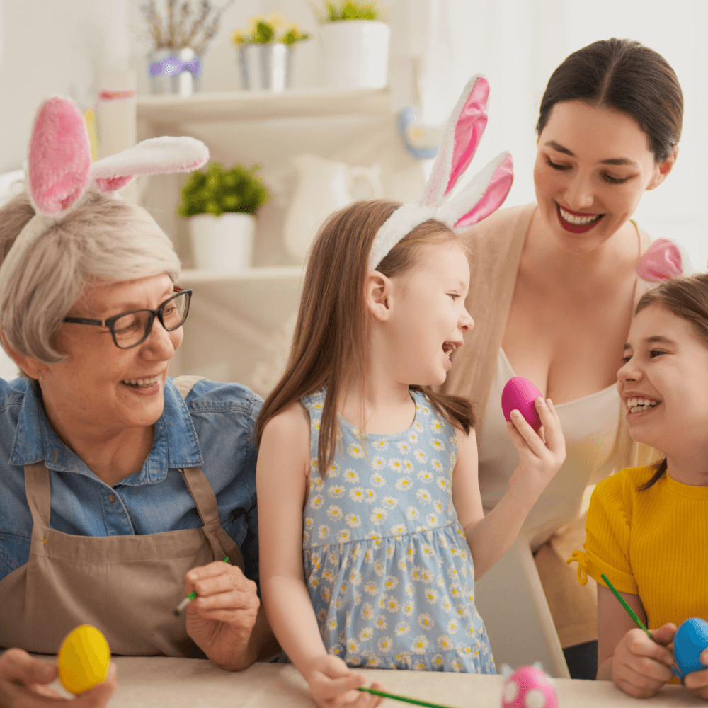 Three generations of women, all wearing bunny ears, joyfully painting Easter eggs together at a decorated table. - Home Instead