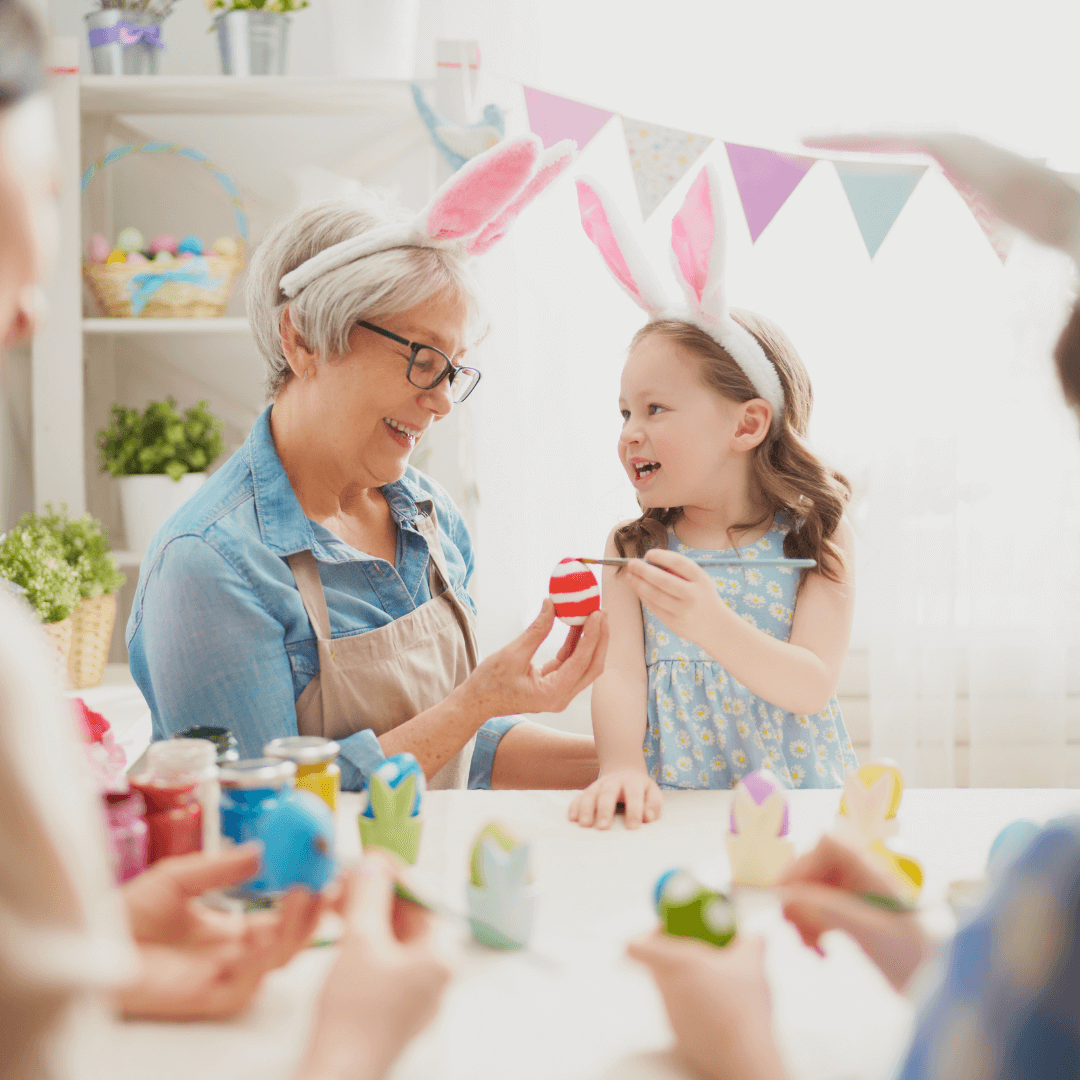 A woman and a girl with bunny ears paint Easter eggs at a table, surrounded by others also painting eggs. - Home Instead