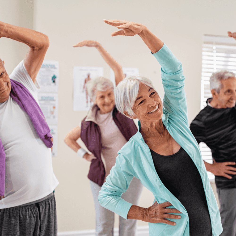Older adults in a fitness class doing side stretches with smiles on their faces, led by a woman in a blue jacket. - Home Instead