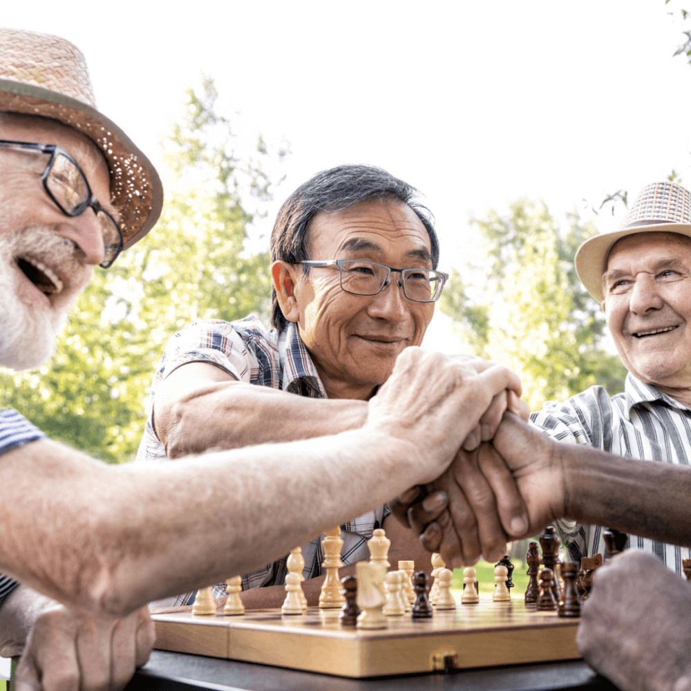 Three elderly men smiling and shaking hands over a chessboard outdoors, enjoying a game together. - Home Instead