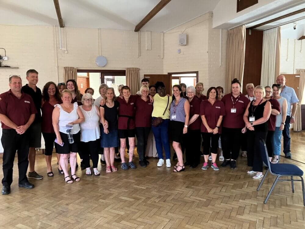 A group of people stand together indoors on a parquet floor, smiling for a photo. - Home Instead