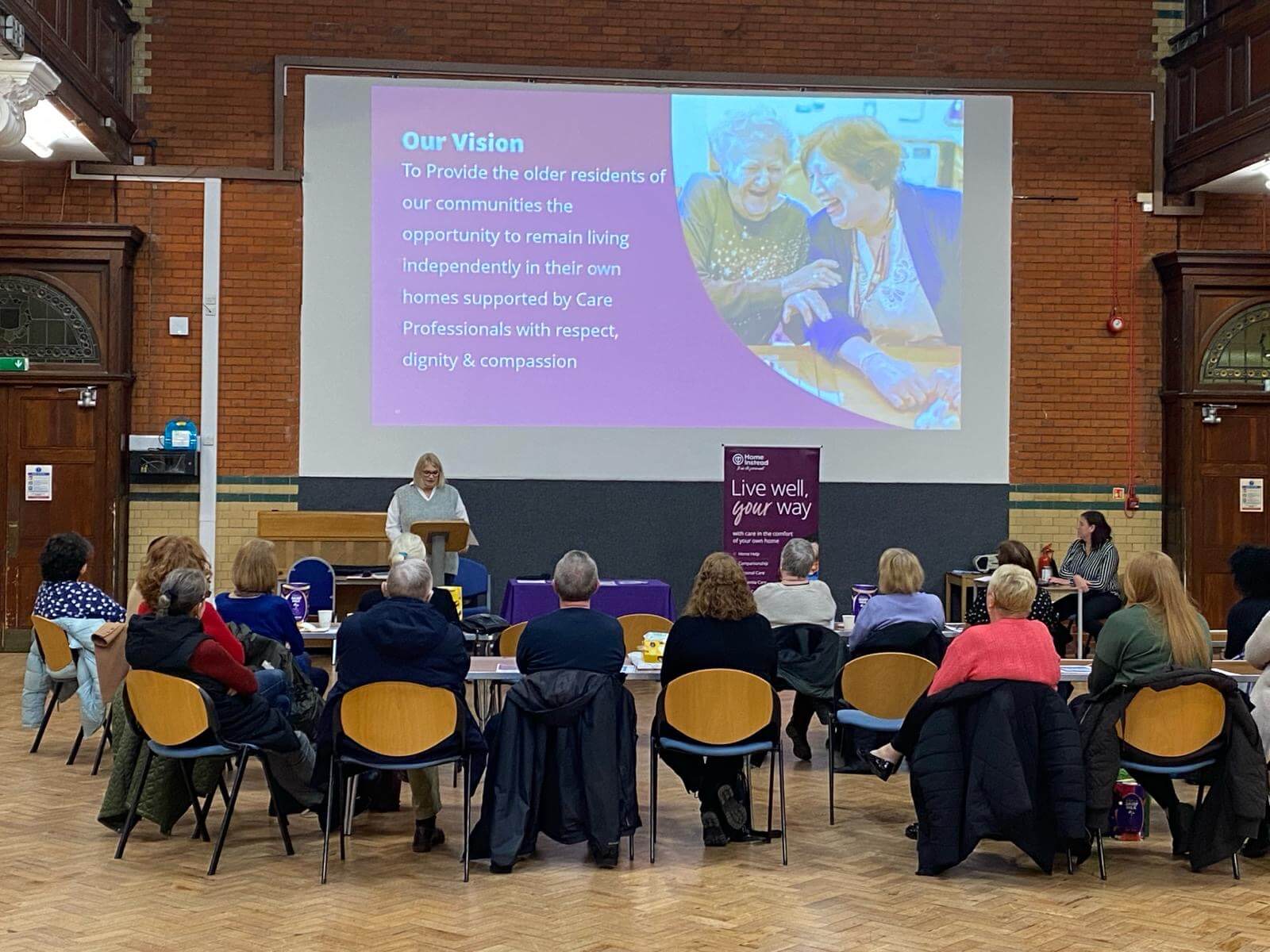 A woman presents to an audience in a hall, with a projection about community care for older residents displayed on-screen. - Home Instead