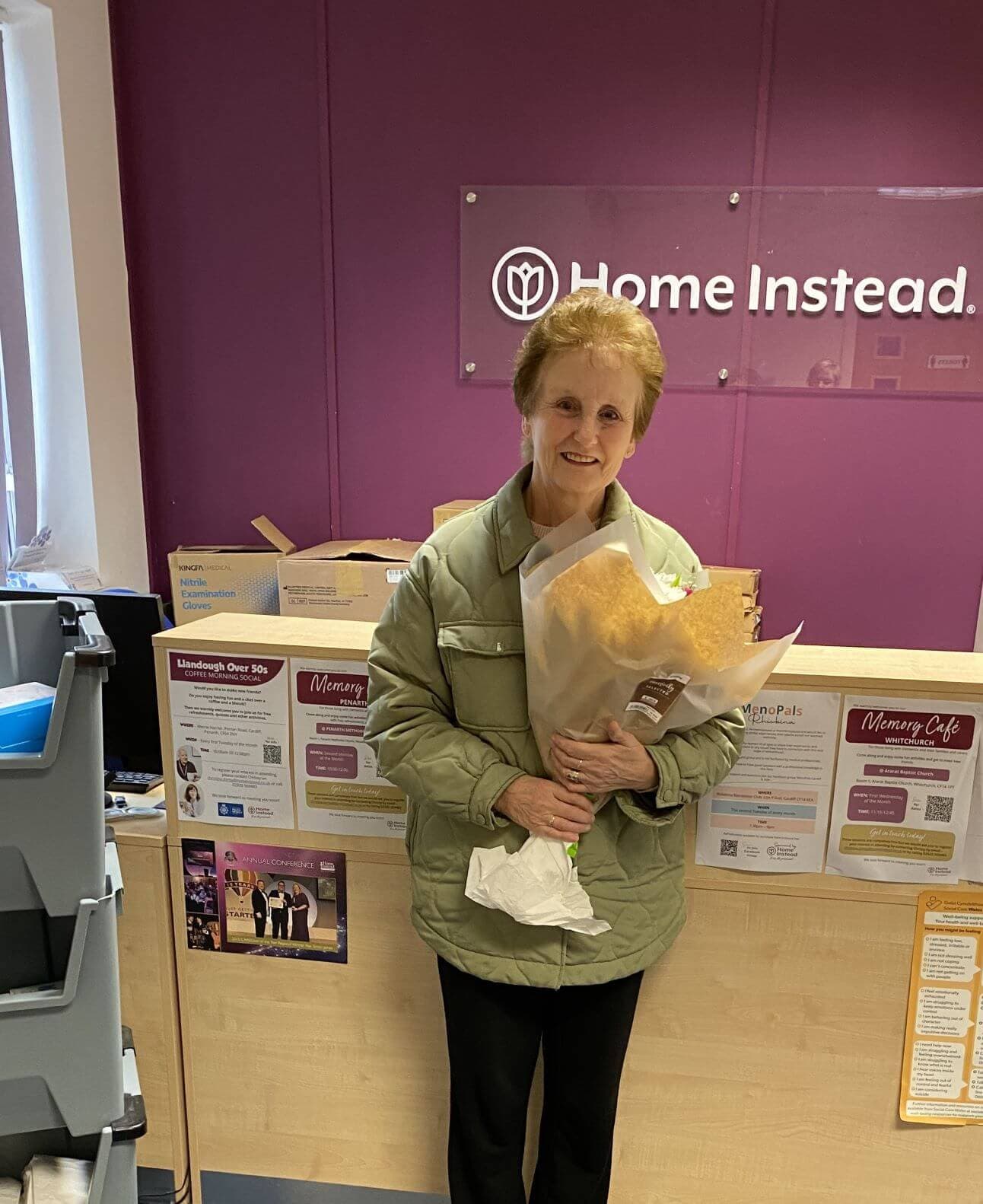 An elderly woman holding a bouquet stands smiling in front of a "Home Instead" sign and informational posters. - Home Instead