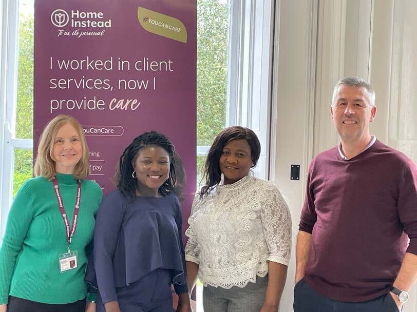 Four smiling people stand indoors next to a sign that says "I worked in client services, now I provide care" at Home Instead. - Home Instead