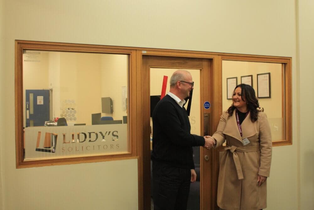 Two people shaking hands in front of an office labeled "Liddy's Solicitors. - Home Instead