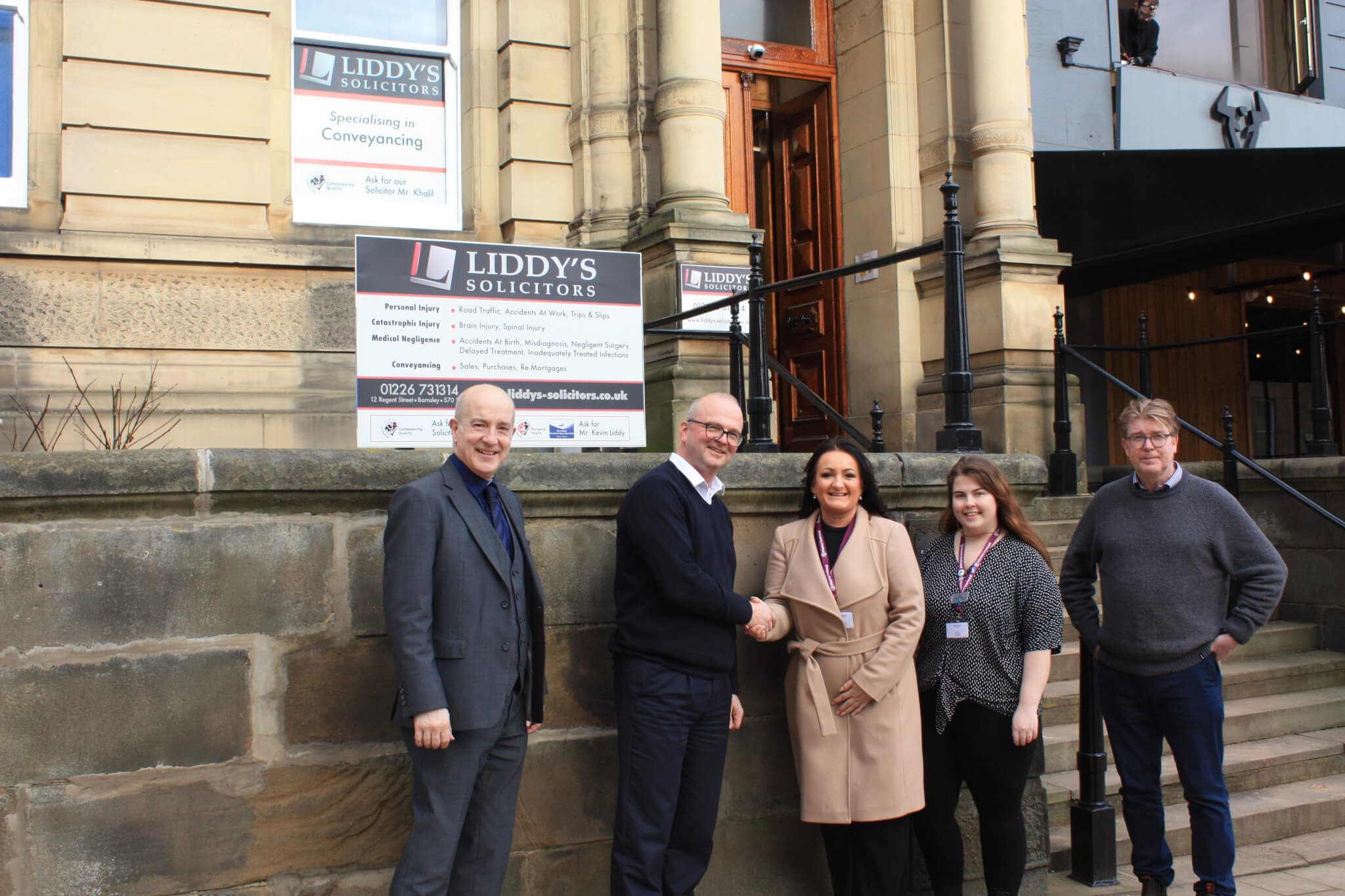 Five people standing in front of a Liddy's Solicitors sign outside a building, with two shaking hands. - Home Instead
