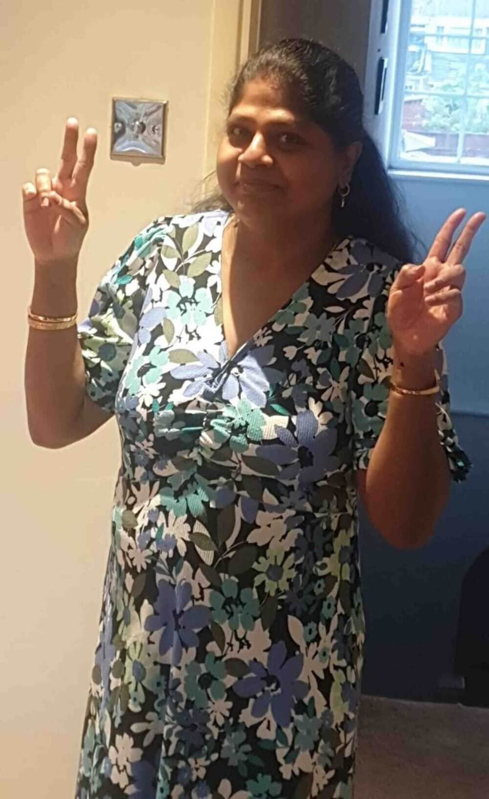 A woman in a floral dress is giving a peace sign with both hands, standing indoors with a cheerful expression. - Home Instead