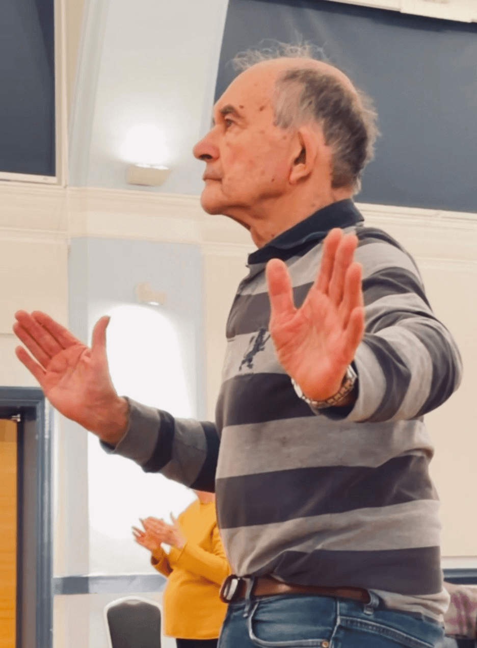 An elderly man stands indoors with his hands raised in front of him, wearing a striped shirt and jeans. - Home Instead