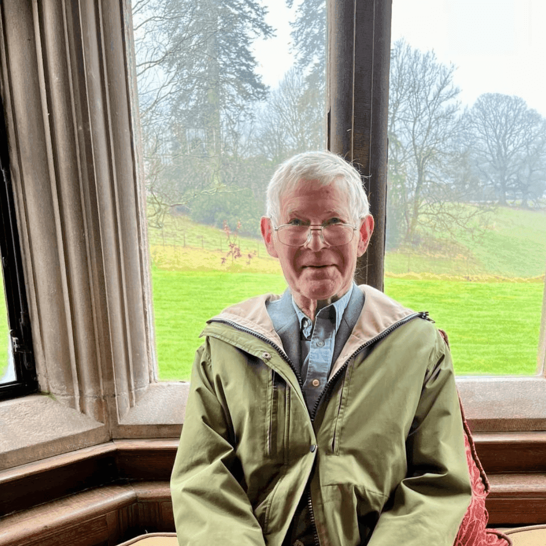 Elderly man in a green jacket sits by a large window with a scenic outdoor view of trees and grassy fields. - Home Instead