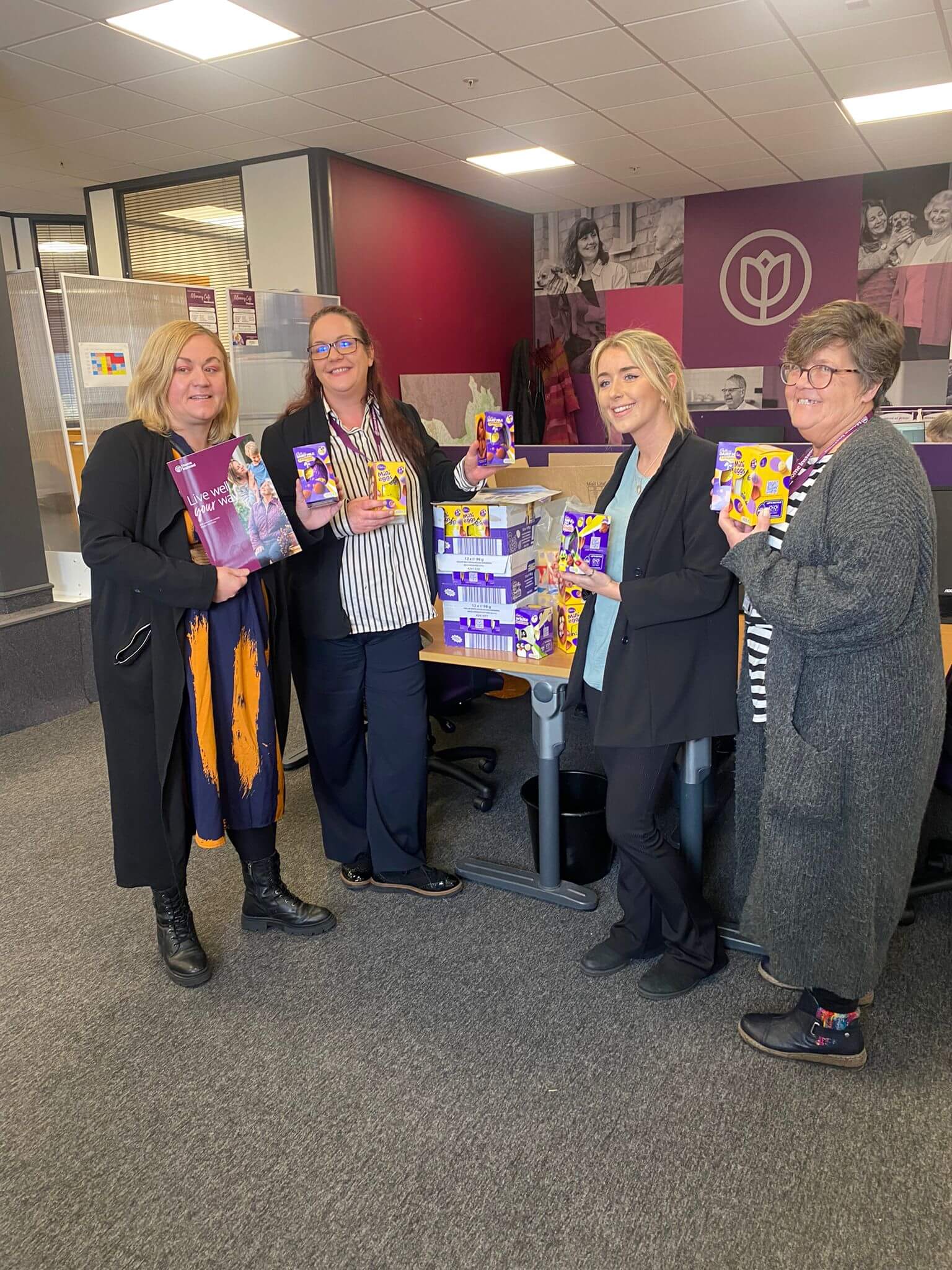 Four women are standing in an office, holding Easter egg boxes and smiling. - Home Instead
