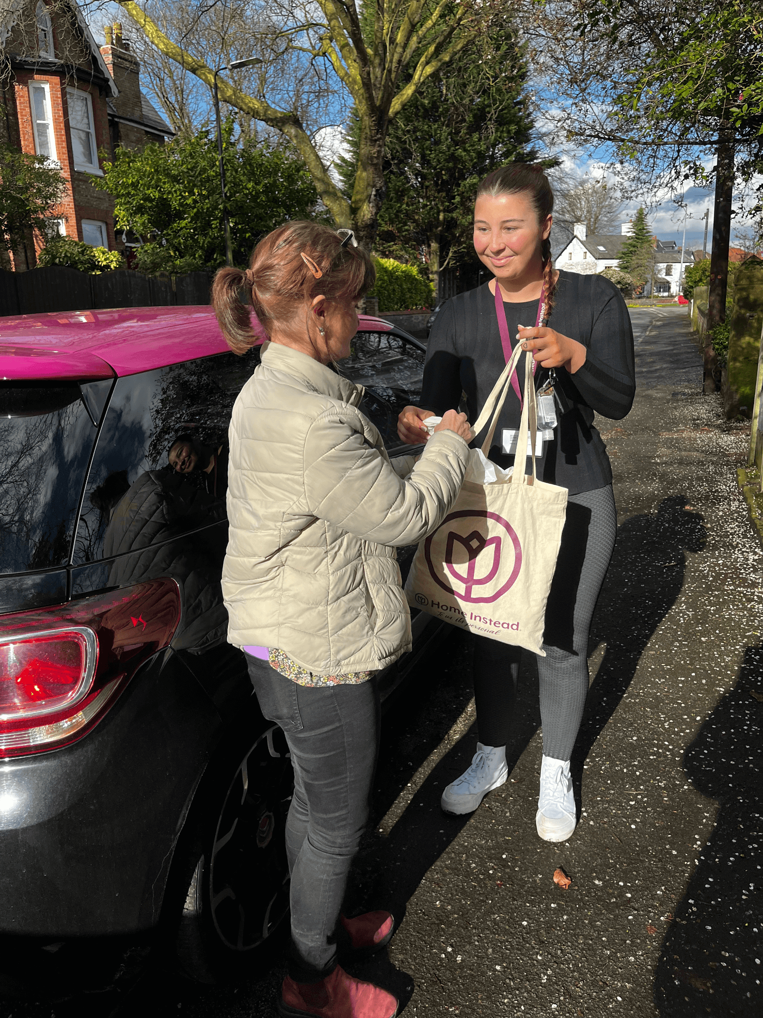 A woman in a gray jacket hands a tote bag to a smiling woman standing by a parked car on a sunny day. - Home Instead