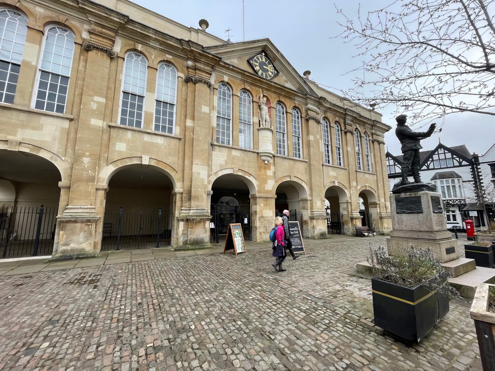 An old building with arched windows and a clock, people walking, and a statue on a cobblestone square under a cloudy sky. - Home Instead