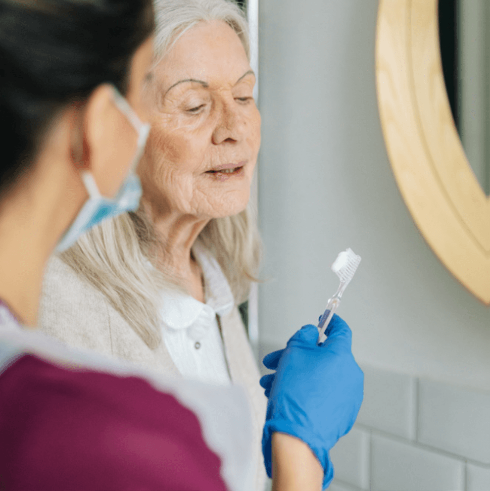 A Home Instead Care Professional with gloves and a mask holds a toothbrush for a female home care client in front of a mirror to assist with personal care.