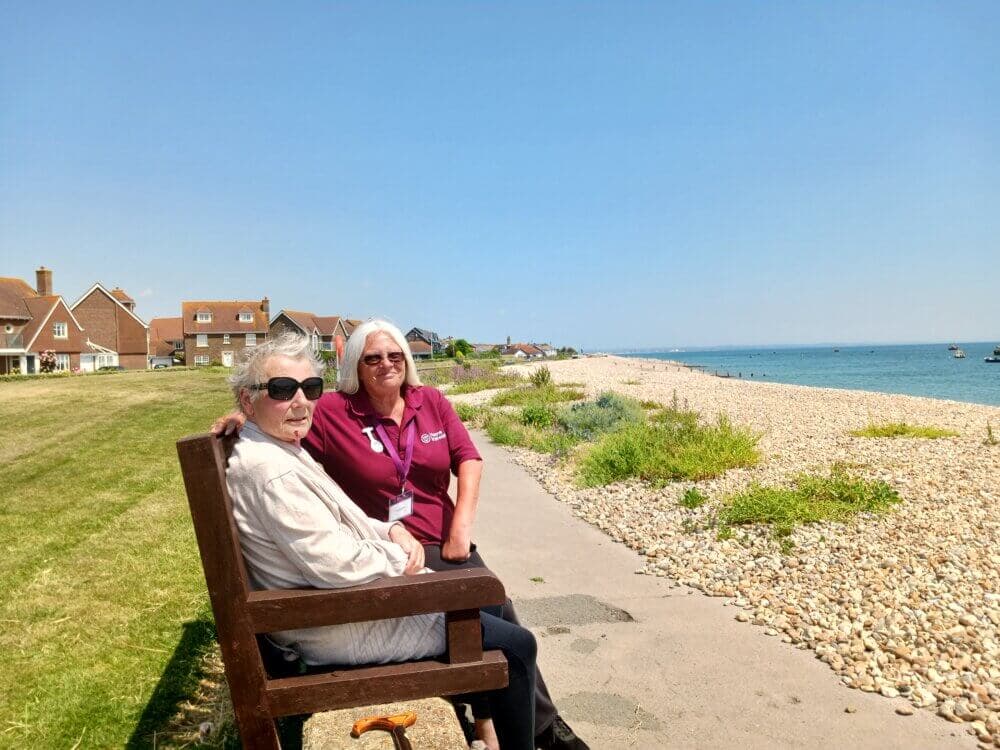 Two women sitting on a bench overlooking a pebble beach with houses in the background on a sunny day. - Home Instead