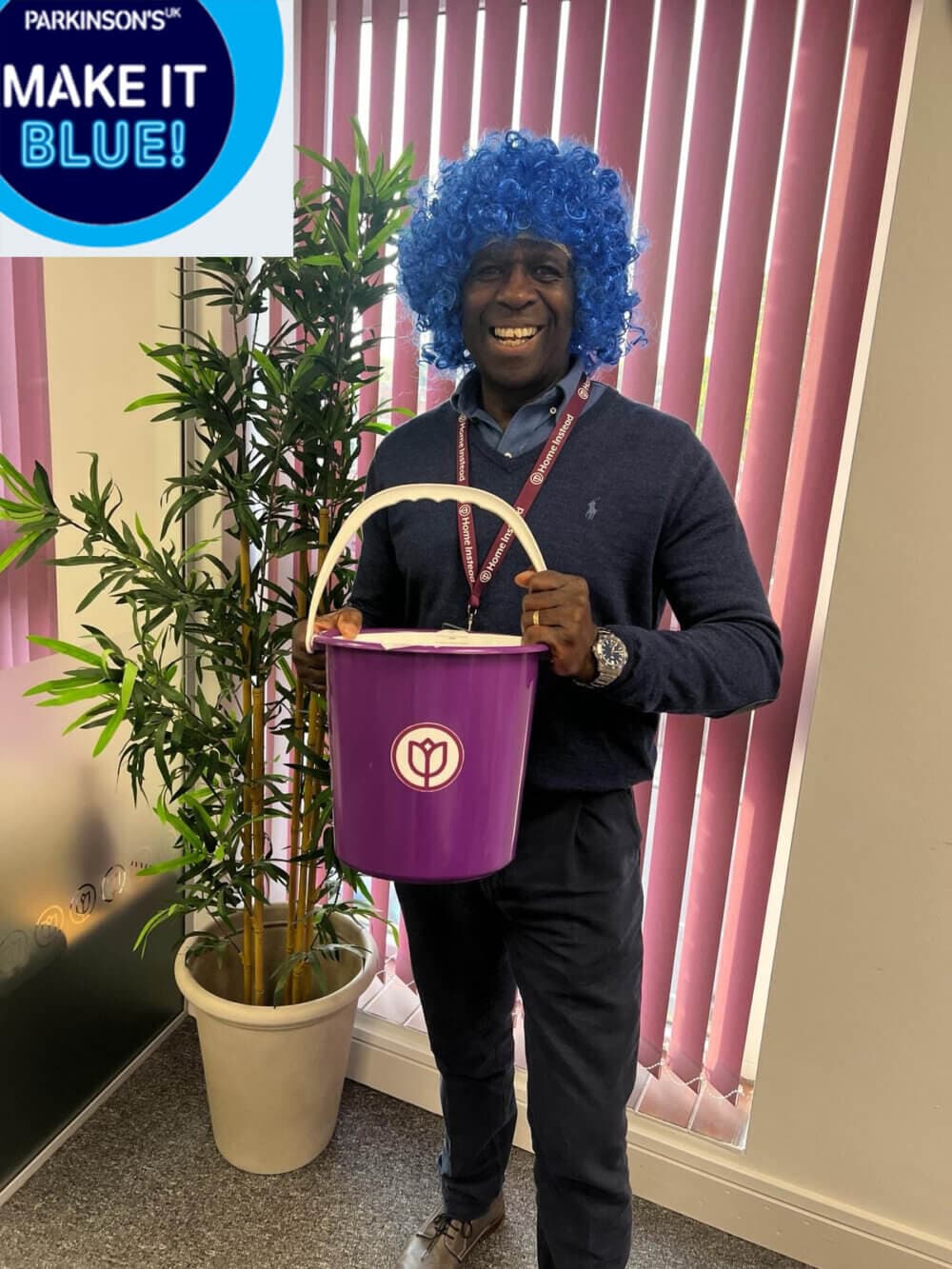 Smiling man with blue wig holding a purple donation bucket, standing next to a plant. "MAKE IT BLUE!" sign above. - Home Instead