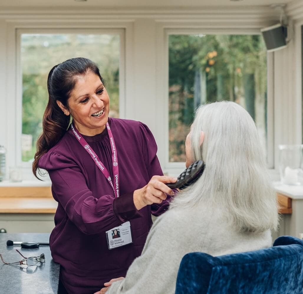 A caregiver smiles while brushing a seated older woman's long white hair in a sunlit room. - Home Instead