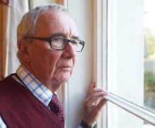 Elderly man in glasses and a red sweater vest looks out of a window with a thoughtful expression. - Home Instead