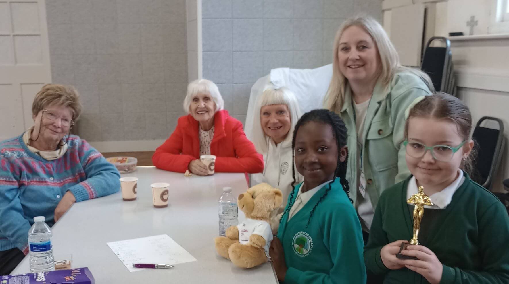 A group of people of various ages sitting at a table, smiling. Two children are holding a teddy bear and a trophy. - Home Instead