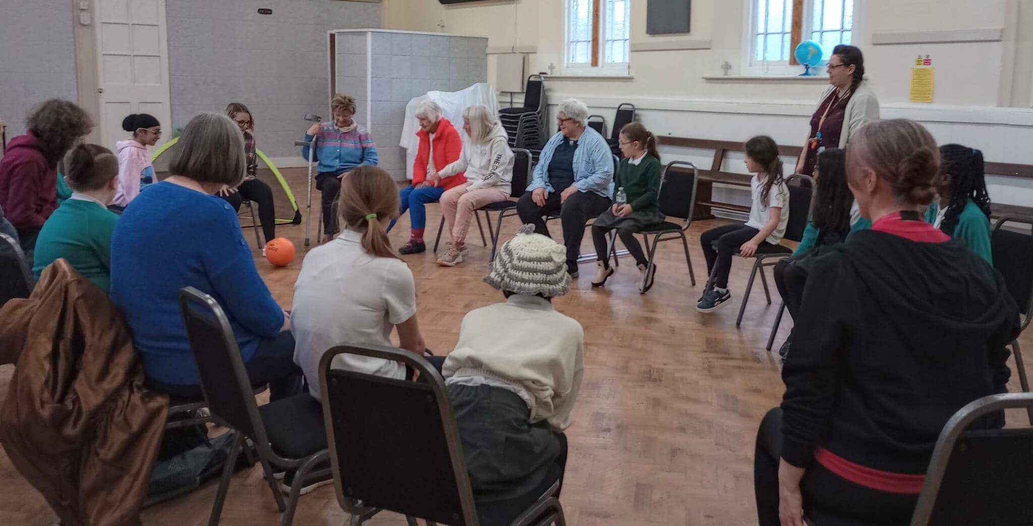 A diverse group of people sit in a circle in a community hall, engaging in a group activity with a ball. - Home Instead