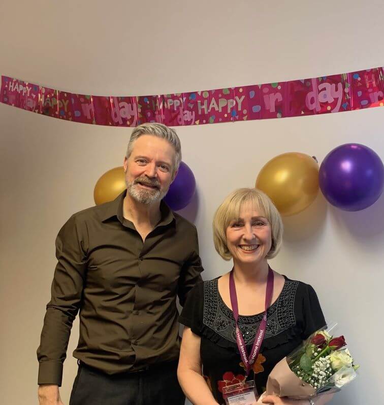 Two people smiling at a birthday party, with balloons and a "Happy Birthday" banner in the background. - Home Instead