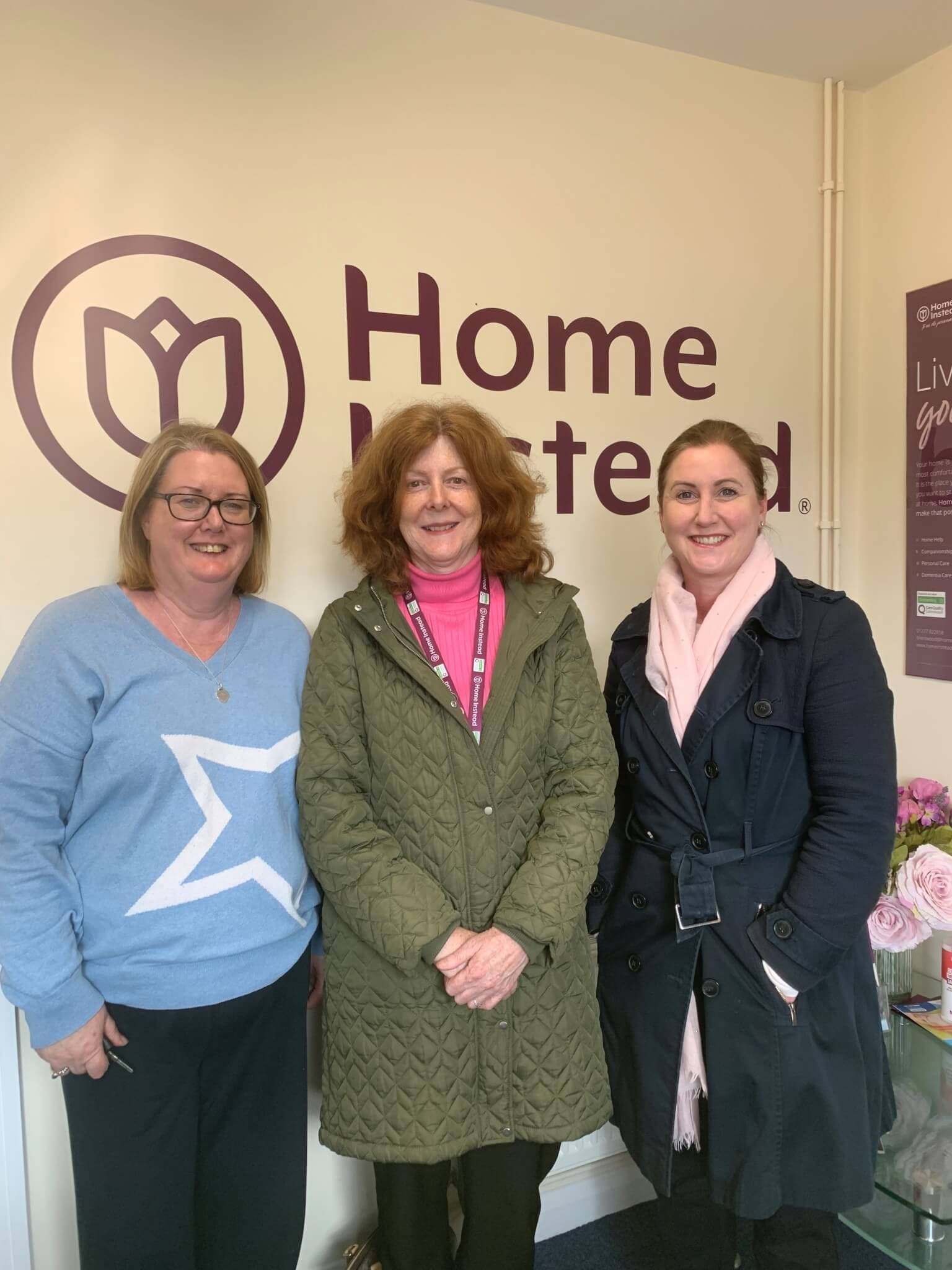 Three women stand smiling in front of a sign that reads "Home Instead" on a wall. They are dressed in casual and business attire. - Home Instead