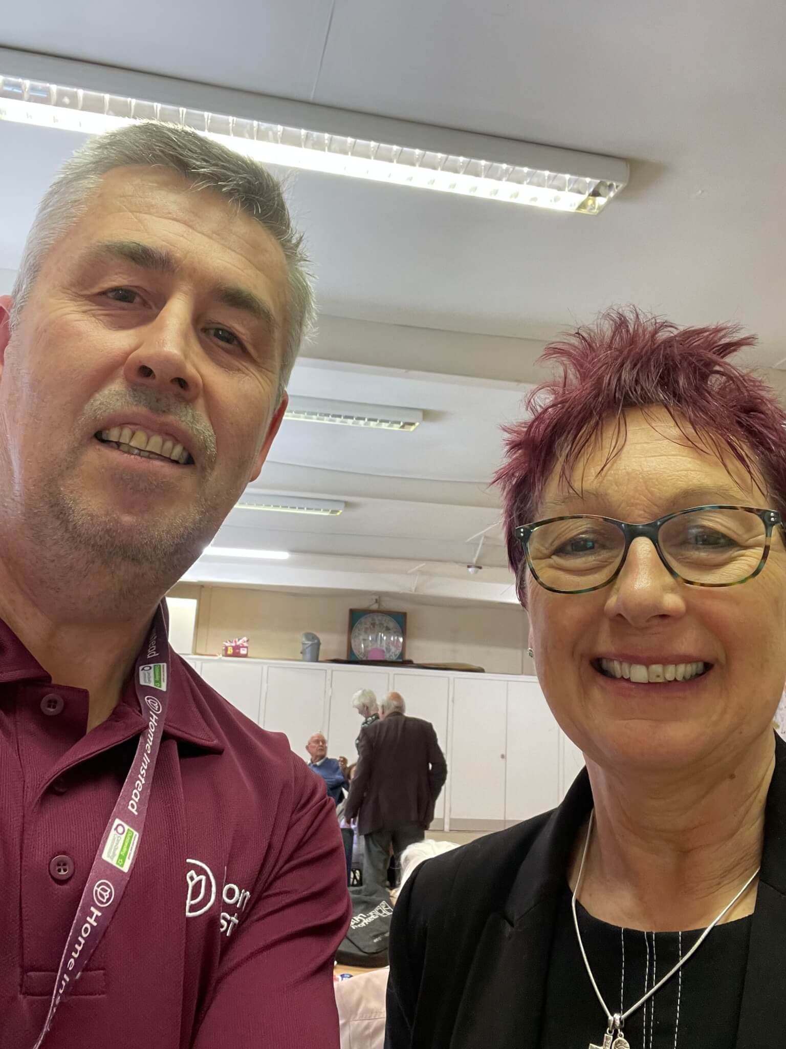 Two people smiling and posing for a selfie indoors, with others seated and conversing in the background. - Home Instead