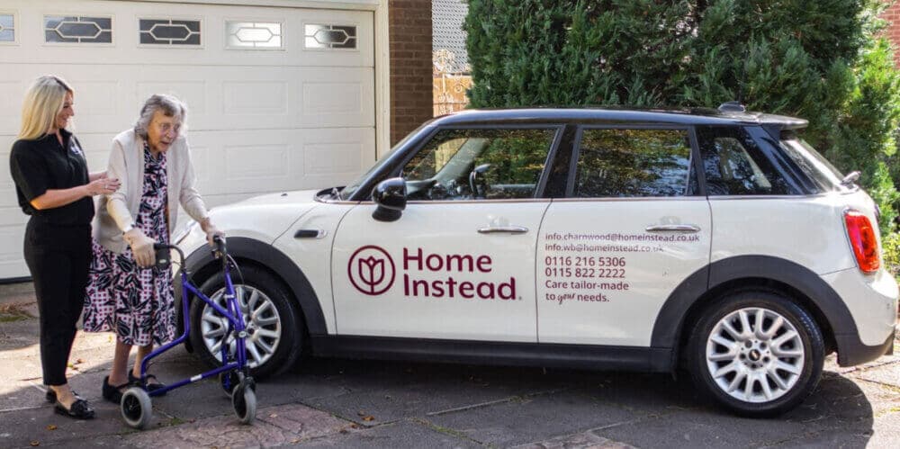 A woman with a walker is assisted by a caregiver beside a car labeled "Home Instead" with contact details. - Home Instead