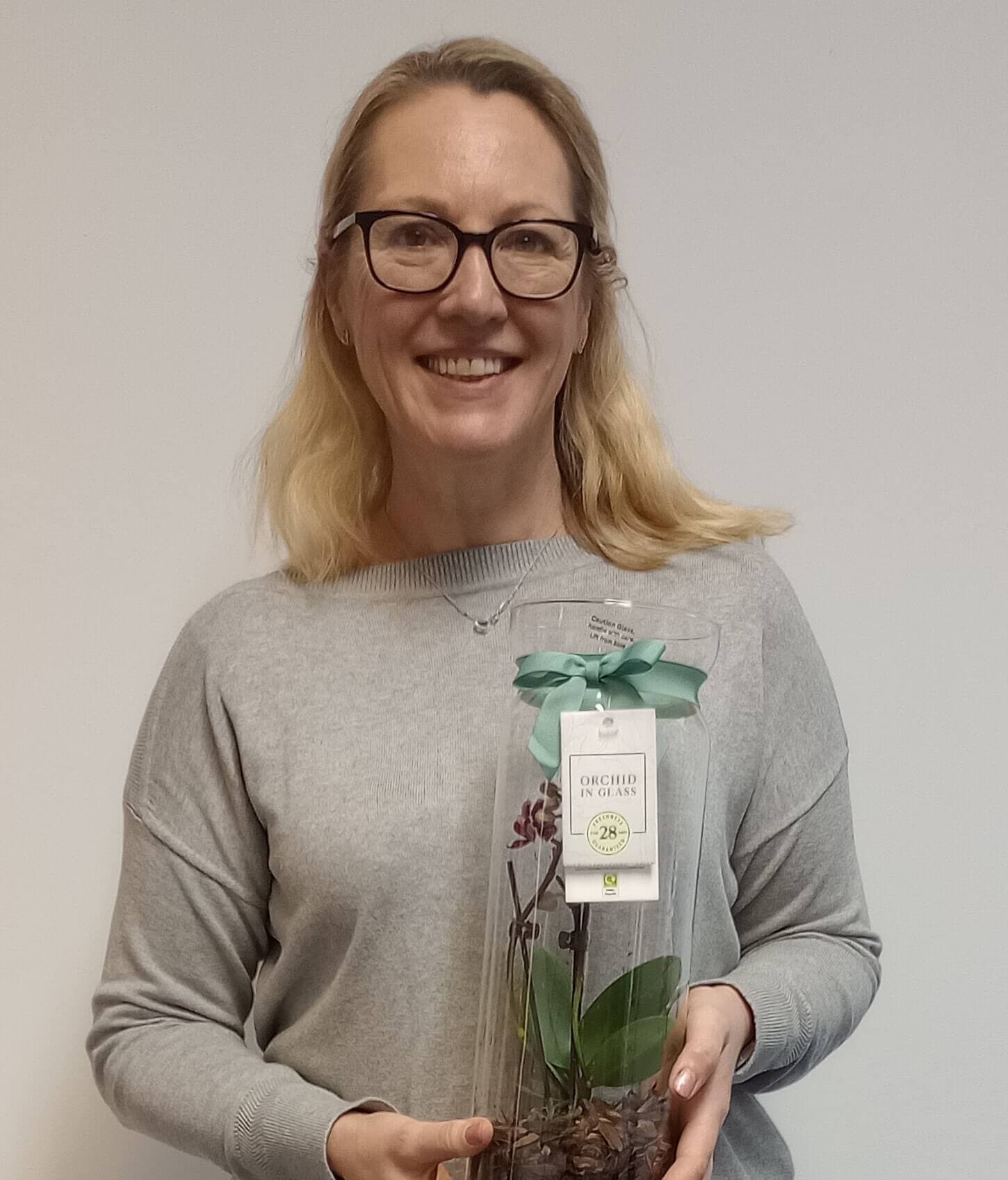 A woman wearing glasses and a grey sweater holds an orchid plant in a clear glass container. - Home Instead