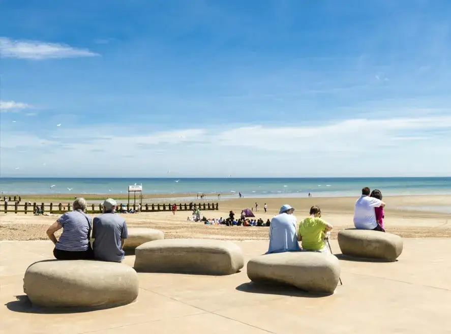 People sitting on large concrete seats by the sea on a sunny day, with a few others on the sandy beach in the background. - Home Instead