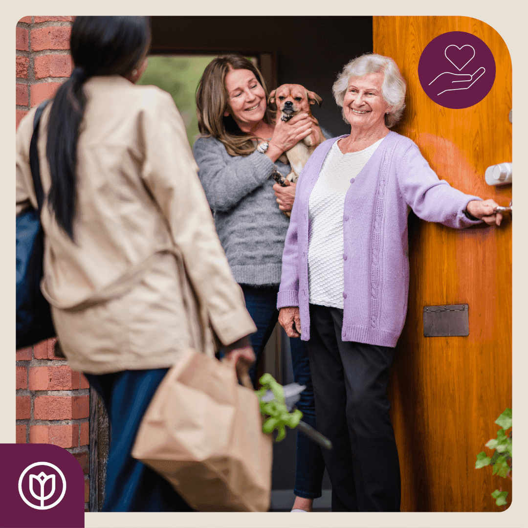 An elderly woman opens the door to a visitor holding a grocery bag, as another woman with a dog stands inside smiling. - Home Instead