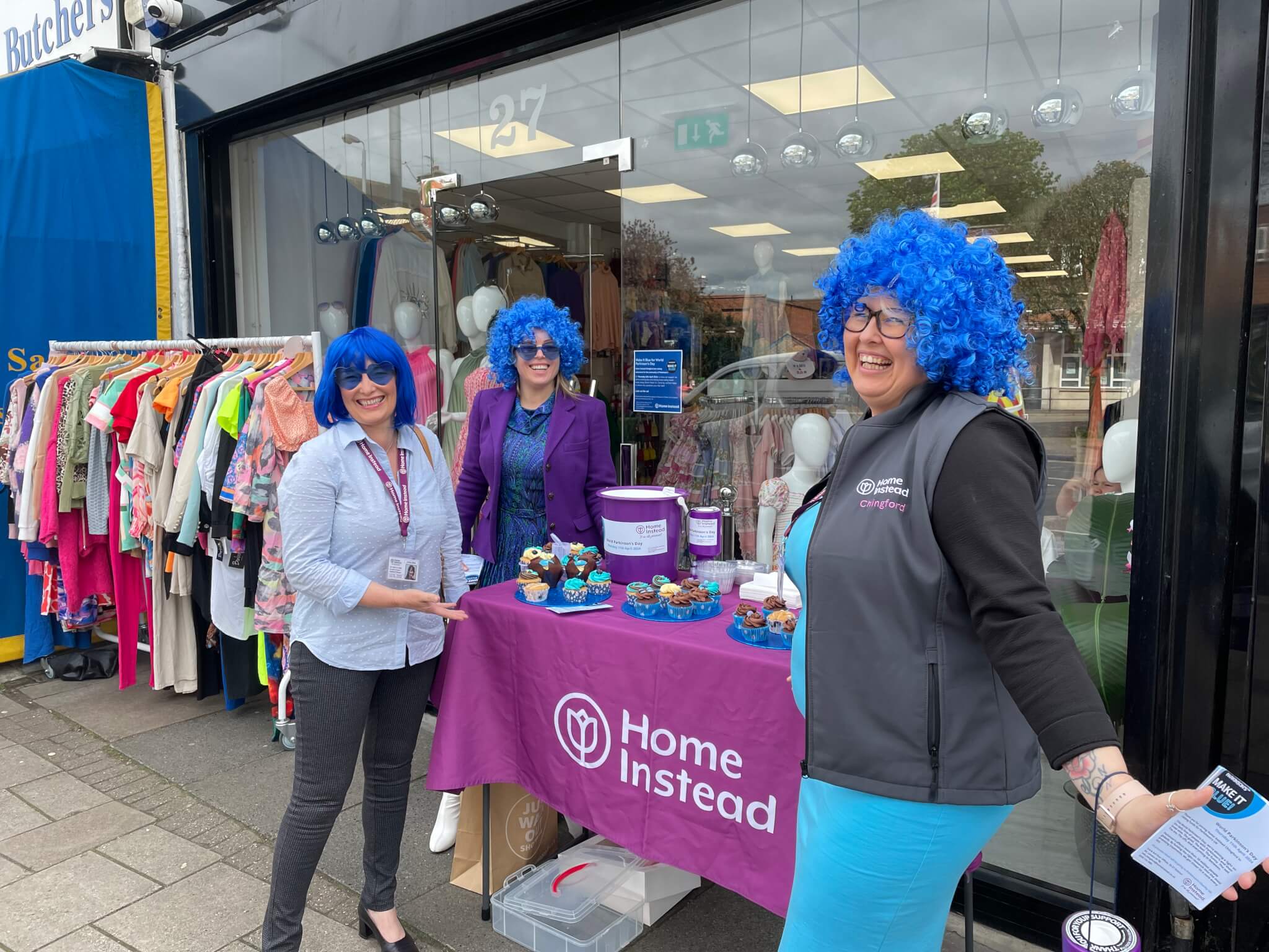 Three people with blue wigs smiling at a table with cupcakes and "Home Instead" banner outside a clothing store. - Home Instead