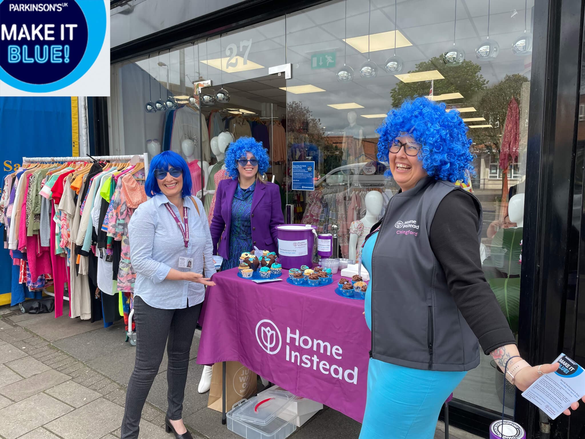 Three women with blue wigs at a charity event table for Home Instead, with clothes displayed behind them. - Home Instead