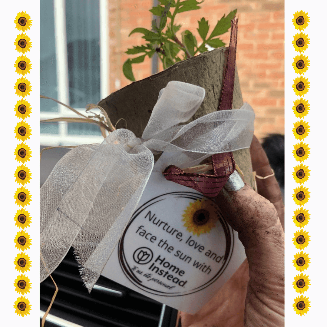 A small plant in a pot with a white ribbon, held by a dirty hand. Text reads "Nurture, love and face the sun with Home Instead". - Home Instead